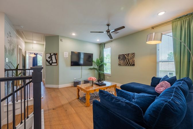 Upstairs loft living room with navy velvet sectional, wooden coffee table with pink flowers, wall-mounted TV, zebra artwork, green curtains and hardwood floors