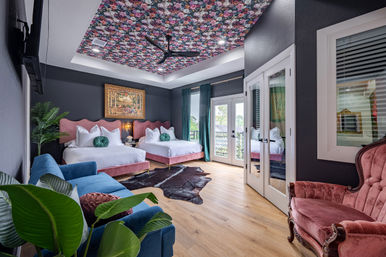 Boutique-style bedroom with floral tray ceiling, dark charcoal walls, two pink velvet beds, teal curtains, blue sofa, vintage pink armchair, French doors to balcony and hardwood floors.