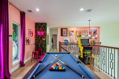 Vibrant loft game room in a home with a blue pool table and crossed cues, Pac‑Man arcade cabinet, pink gallery wall, neon 'Right Place Right Time' greenery and magenta curtains.