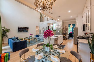 Bright open-concept luxury living room and kitchen with two-story ceiling, ornate chandelier, blue velvet sofa, marble island, glass dining table with pink orchids, hardwood floors and decorative wall mural.