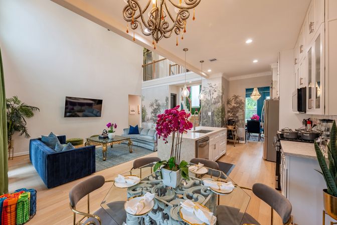 Bright open-concept luxury living room and kitchen with two-story ceiling, ornate chandelier, blue velvet sofa, marble island, glass dining table with pink orchids, hardwood floors and decorative wall mural.