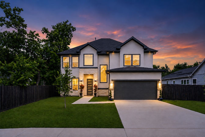 Twilight view of a modern two-story suburban house with warm interior lights, attached two-car garage, clean concrete driveway, manicured front lawn, potted planters by the dark front door and a colorful orange-purple sunset sky.