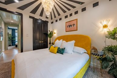 Modern bedroom with mustard yellow velvet bed and white linens, gold sunburst chandelier and wall sconces, black double doors and black-and-white striped ceiling rays, tropical plants and a small gold side table