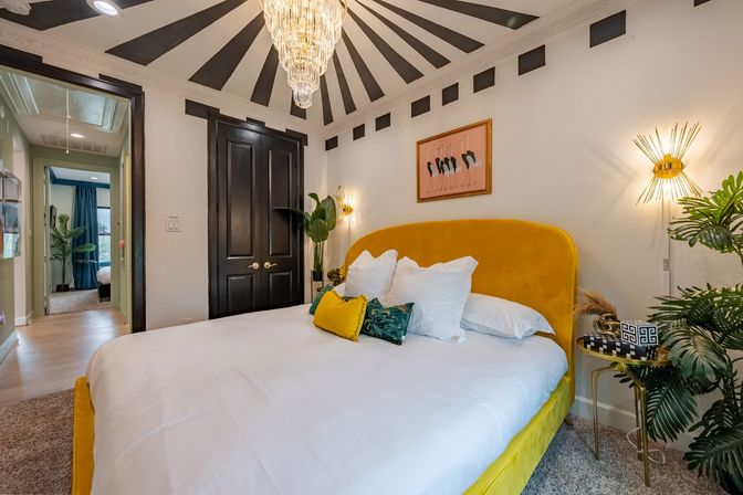 Modern bedroom with mustard yellow velvet bed and white linens, gold sunburst chandelier and wall sconces, black double doors and black-and-white striped ceiling rays, tropical plants and a small gold side table