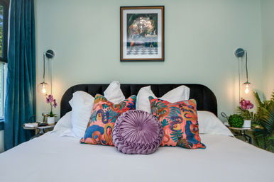 Bright modern bedroom with white bedding, black velvet headboard, vibrant tropical-print throw pillows and a round mauve velvet cushion, teal curtains, hanging globe bedside lamps and potted orchids