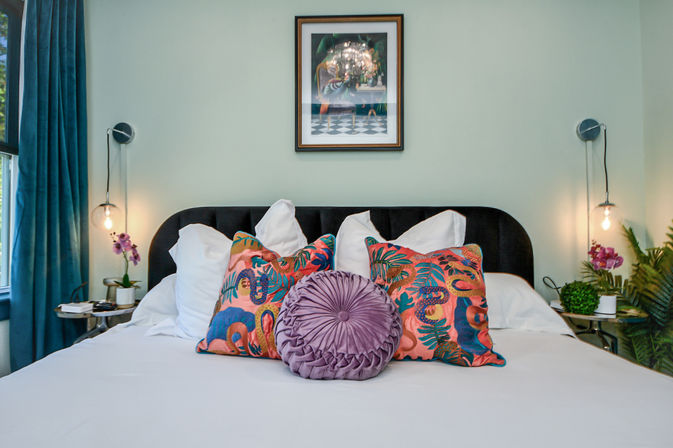 Bright modern bedroom with white bedding, black velvet headboard, vibrant tropical-print throw pillows and a round mauve velvet cushion, teal curtains, hanging globe bedside lamps and potted orchids