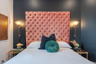 Stylish bedroom interior with a plush pink tufted velvet headboard against a dark gray accent wall, brass wall sconces, white bedding and layered decorative pillows on gold mirrored bedside tables with flowers and books.