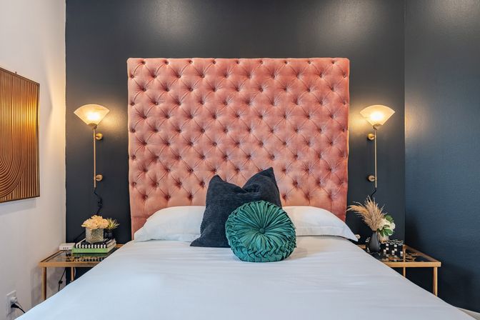 Stylish bedroom interior with a plush pink tufted velvet headboard against a dark gray accent wall, brass wall sconces, white bedding and layered decorative pillows on gold mirrored bedside tables with flowers and books.