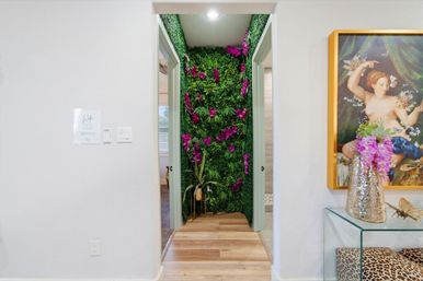 Modern home hallway with an indoor vertical green wall and bright purple orchids in a recessed nook, wood-plank floors, framed classical painting and metallic vase with pink blooms.