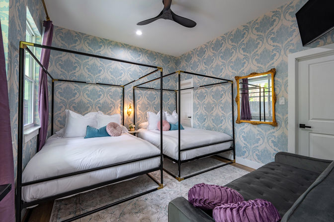 Chic guest bedroom with two black metal canopy beds dressed in white linens and pink and teal accent pillows, blue damask wallpaper, gold-framed mirror, purple curtains, velvet gray sofa with round purple cushions, and black ceiling fan.