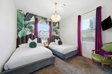 Sunlit guest bedroom with two twin beds, gray velvet scalloped headboards, white linens and round green accent pillows, bold banana-leaf tropical accent wall, magenta floor-length curtains, bubble chandelier and a vintage green upholstered armchair.