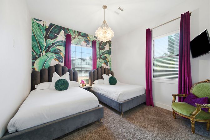 Sunlit guest bedroom with two twin beds, gray velvet scalloped headboards, white linens and round green accent pillows, bold banana-leaf tropical accent wall, magenta floor-length curtains, bubble chandelier and a vintage green upholstered armchair.