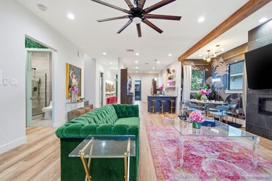 Open-concept modern living room and kitchen with green velvet sofa, vibrant pink patterned rug, glass coffee table, island with brass bar stools, tropical wallpapered dining area, exposed wood beam, oversized ceiling fan, and light hardwood floors.