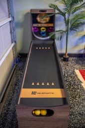 Tabletop skee-ball arcade game in a home game room — wood-framed lane with three yellow balls in the front, colorful target rings and a potted palm nearby.
