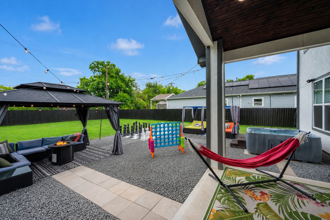 Family-friendly suburban backyard patio and outdoor living space with covered lounge and fire pit under string lights, red hammock on tiled porch, giant Connect Four and oversized chess set on gravel, hot tub and fenced green lawn.