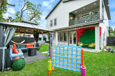 Modern suburban two-story house backyard with covered lounge gazebo lit by string lights, blue sectional and fire pit, hammock, giant Connect Four on green lawn, decorative vertical green wall with red flowers and yellow patio chairs.