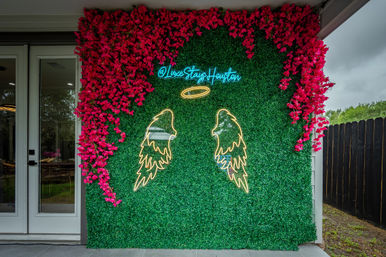 Neon angel wings and halo on a green boxwood photo wall framed by bright pink flowers outside a modern Houston home — trendy outdoor photo backdrop.