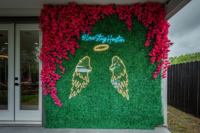 Neon angel wings and halo on a green boxwood photo wall framed by bright pink flowers outside a modern Houston home — trendy outdoor photo backdrop.