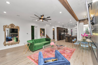 Bright open-concept modern home interior with a green velvet tufted sofa, blue velvet chair, pink patterned rug, ornate gold wall mirror, glass coffee table, wood floors, exposed beam and large ceiling fan overlooking a sleek kitchen and gold-accent dining area.