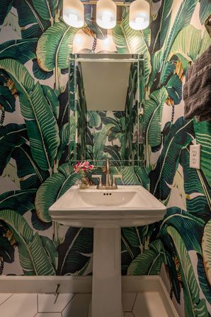 Powder room with bold green banana‑leaf wallpaper, white pedestal sink and brushed brass faucet, mirrored medicine cabinet, three globe vanity lights and small pink flowers in a glass vase.