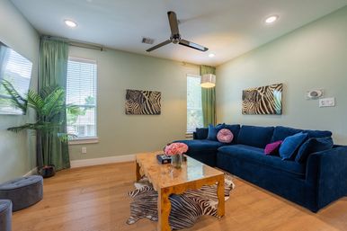 Sunlit modern living room with a navy velvet sectional, colorful throw pillows, wooden coffee table with pink roses, zebra-print rug and wall art, green curtains, hardwood floors and ceiling fan