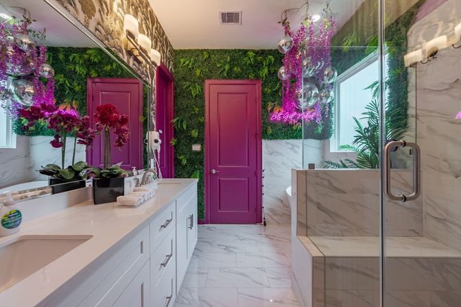 Vibrant luxury bathroom with marble floors and glass shower, white double vanity and large mirror, magenta door, lush green living wall with neon pink sign, hanging disco balls and purple orchids.