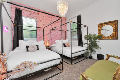 Modern glam bedroom with two black four-poster twin beds dressed in white linens against a pink flamingo wallpaper accent wall, crystal chandelier overhead, gold ornate mirror, potted palm, dark curtains, and green velvet bench.
