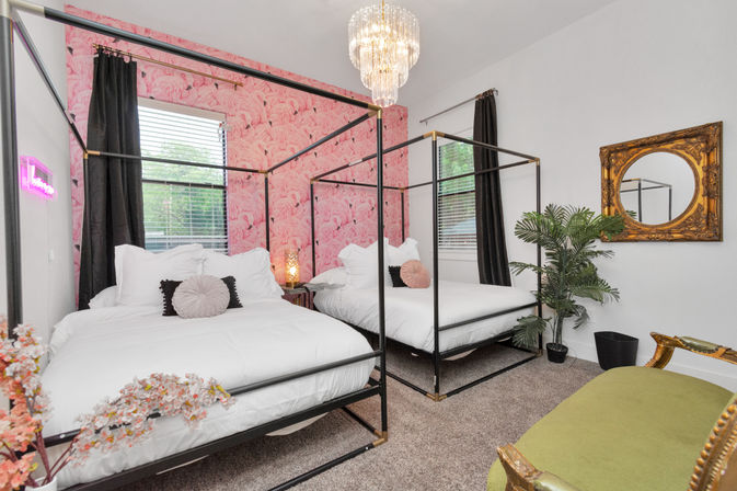 Modern glam bedroom with two black four-poster twin beds dressed in white linens against a pink flamingo wallpaper accent wall, crystal chandelier overhead, gold ornate mirror, potted palm, dark curtains, and green velvet bench.