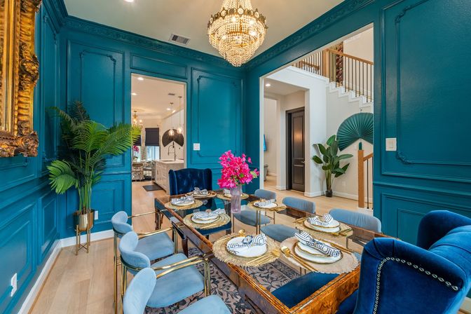 Teal-paneled dining room with crystal chandelier, glass-top table set for six, blue velvet chairs, bright pink floral centerpiece and open view to kitchen and staircase.