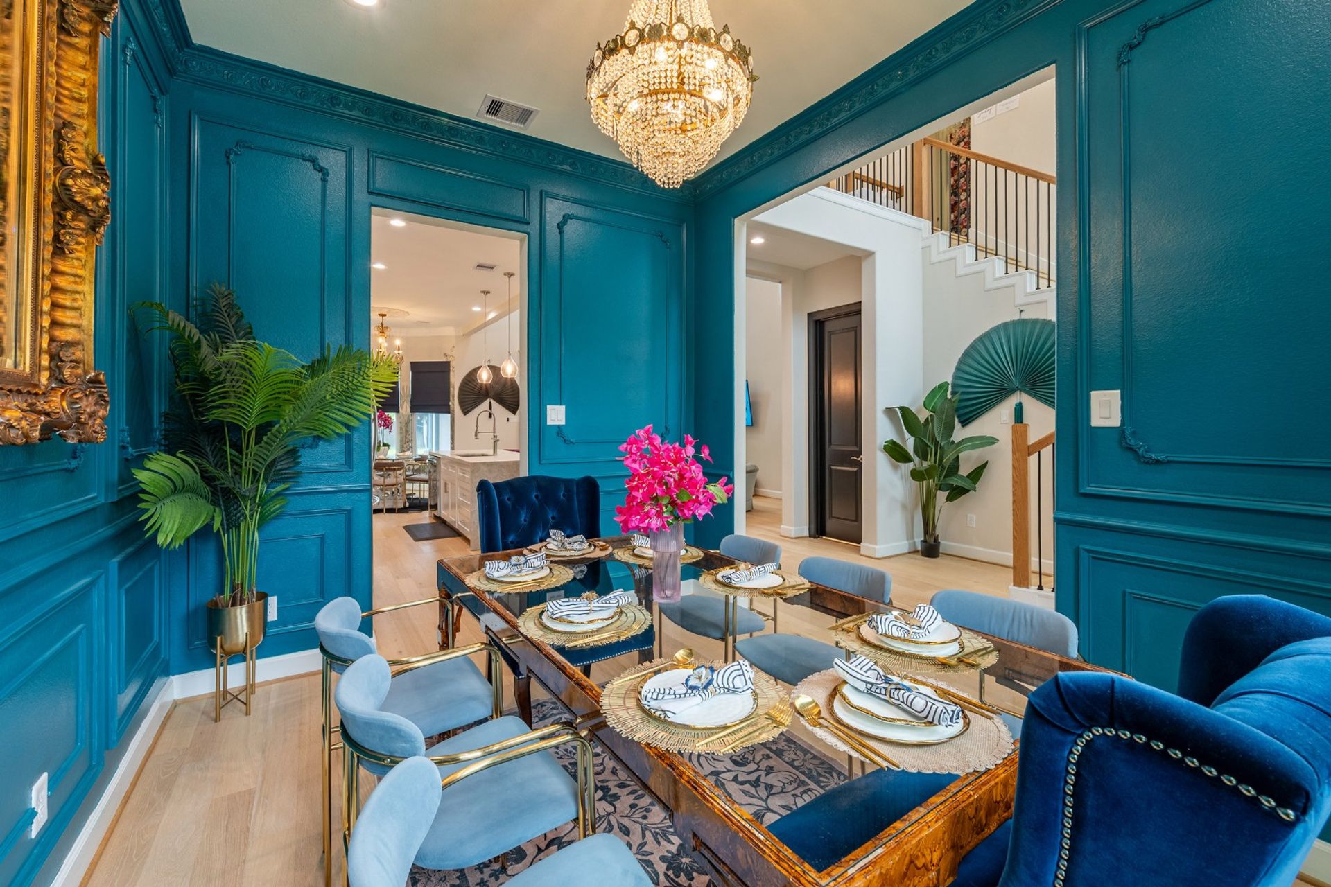 Teal-paneled dining room with crystal chandelier, glass-top table set for six, blue velvet chairs, bright pink floral centerpiece and open view to kitchen and staircase.
