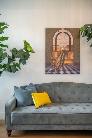 Cozy modern living room interior with a gray tufted velvet sofa, pop-of-yellow accent pillow, leafy houseplants, and wall art featuring a peacock under a chandelier.