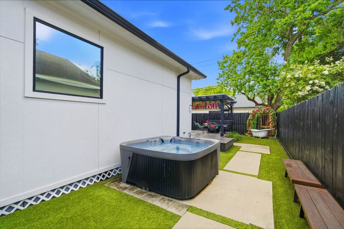 Backyard patio with a square hot tub on artificial turf, concrete stepping stones leading to a pergola dining area and a flower‑decorated clawfoot tub beside a tall wooden fence.