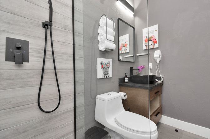 Modern compact bathroom with glass shower and gray wood-look tile, matte black shower fixtures, white toilet, wood vanity with black vessel sink and purple orchid, stacked white towels, floral wall art and wall-mounted hair dryer.