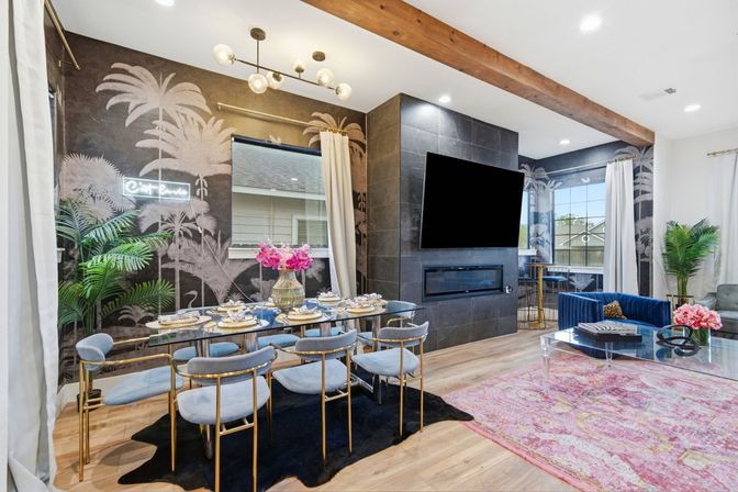 Modern open-concept living and dining room with tropical palm mural, glass dining table, blue velvet chairs, wall-mounted TV above linear fireplace, exposed wood beam and pink area rug.