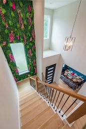 Overhead view of a modern light-wood staircase in a two-story foyer featuring a tall green living wall with pink flowers, a black contemporary front door, hanging pendant chandelier and a bright dining area set below.