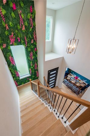 Overhead view of a modern light-wood staircase in a two-story foyer featuring a tall green living wall with pink flowers, a black contemporary front door, hanging pendant chandelier and a bright dining area set below.