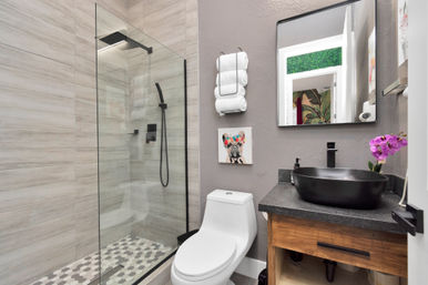 Contemporary bathroom interior with glass walk-in shower and black rain showerhead, hexagon tile floor, white toilet, black vessel sink on wooden vanity, rolled towels and purple orchid.