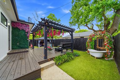 Backyard deck with a dark wooden pergola draped in hanging pink flowers and string lights, bench swing and lounge chairs on a raised deck, faux-grass lawn, decorative clawfoot tub beneath a flower-covered arch by a large tree, and a green floral wall by the house.