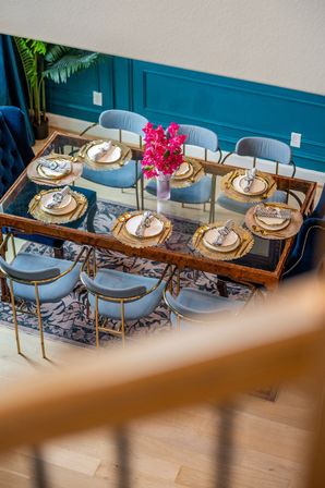 Modern residential dining room with glass-top table set for six — gold chargers, zebra-striped napkins, bright pink floral centerpiece, powder-blue velvet chairs, teal paneled wall and patterned rug.