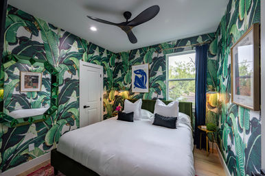 Vibrant tropical bedroom featuring banana-leaf wallpaper, green velvet headboard, crisp white bedding with black accent pillows, geometric green mirror, gold bedside table and lamp, and a modern black ceiling fan by a window with navy curtain.