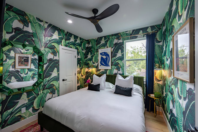 Vibrant tropical bedroom featuring banana-leaf wallpaper, green velvet headboard, crisp white bedding with black accent pillows, geometric green mirror, gold bedside table and lamp, and a modern black ceiling fan by a window with navy curtain.