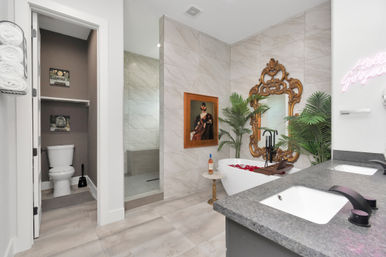 Spa-like modern luxury bathroom with marble tile, freestanding tub with rose petals and wooden tray, ornate gold mirror, double vanity, glass shower, potted tropical plants, and separate toilet niche.