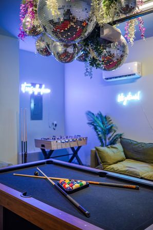 Party-ready indoor game room with black pool table and racked billiard balls, crossed cues on the table, mirrored disco balls and hanging greenery above, neon wall signs, foosball table and a cozy sofa with potted plant in the background.
