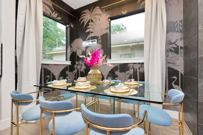Modern dining nook with glass table, gold-accented blue velvet chairs, tropical palm mural wallpaper, vibrant pink orchid centerpiece and neatly set place settings beneath corner windows