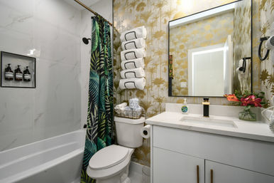 Modern tropical-themed bathroom interior with white tub and marble-look tile, green palm-print shower curtain, gold palm wallpaper, rolled towels above the toilet, and a white vanity with black-and-gold faucet and mirror.