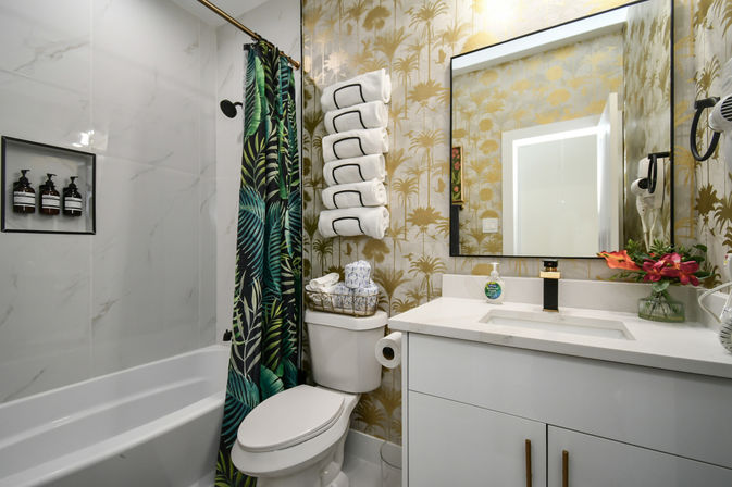 Modern tropical-themed bathroom interior with white tub and marble-look tile, green palm-print shower curtain, gold palm wallpaper, rolled towels above the toilet, and a white vanity with black-and-gold faucet and mirror.