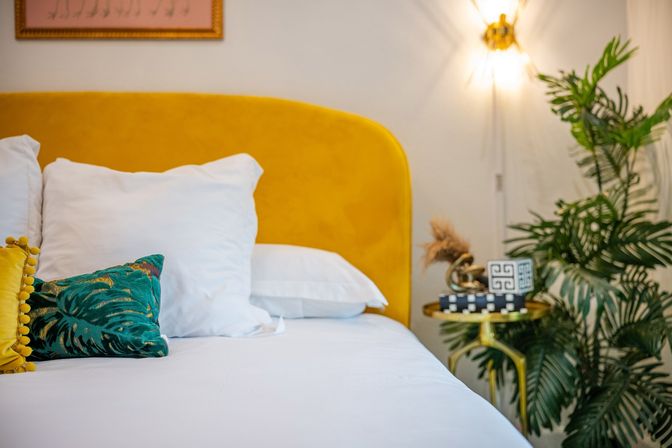 Sunny modern bedroom interior with mustard-yellow velvet headboard, crisp white bedding, teal tropical throw pillow, gold bedside table with decorative accents, glowing wall sconce and lush potted palm.
