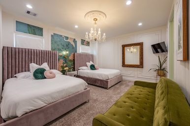 Cozy luxury guest room with two mauve velvet beds, botanical mural accent wall, crystal chandelier, gold-framed mirror, wall-mounted TV and green velvet sofa