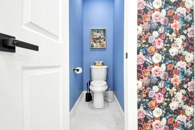 Small modern powder room with bright blue walls and white toilet on marble-look tile, gold toilet paper holder, black brush set and clear waste bin, framed vintage swimsuit print above the tank and bold floral wallpaper visible in the adjacent hallway.