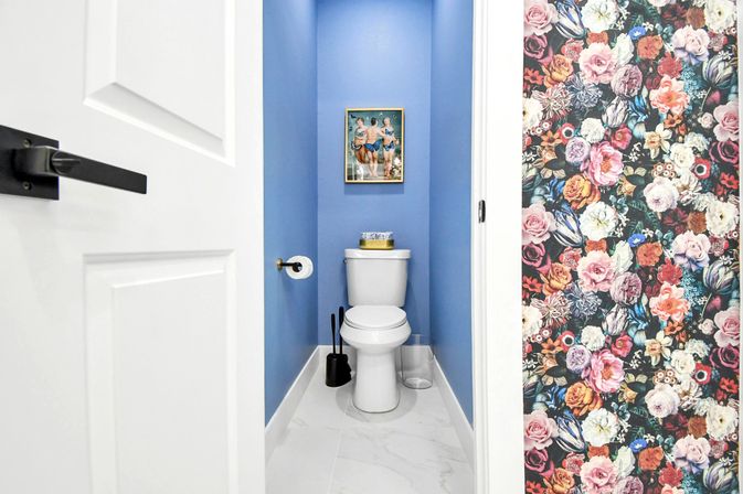 Small modern powder room with bright blue walls and white toilet on marble-look tile, gold toilet paper holder, black brush set and clear waste bin, framed vintage swimsuit print above the tank and bold floral wallpaper visible in the adjacent hallway.